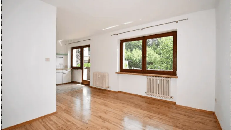 Helles Wohnzimmer mit Holzboden, weißen Wänden und Blick ins Grüne, angrenzend an eine Küche.