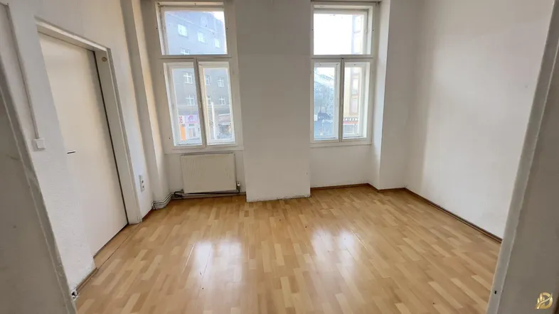 Leeres Zimmer mit Parkettboden und zwei großen Fenstern, die viel Tageslicht hereinlassen.