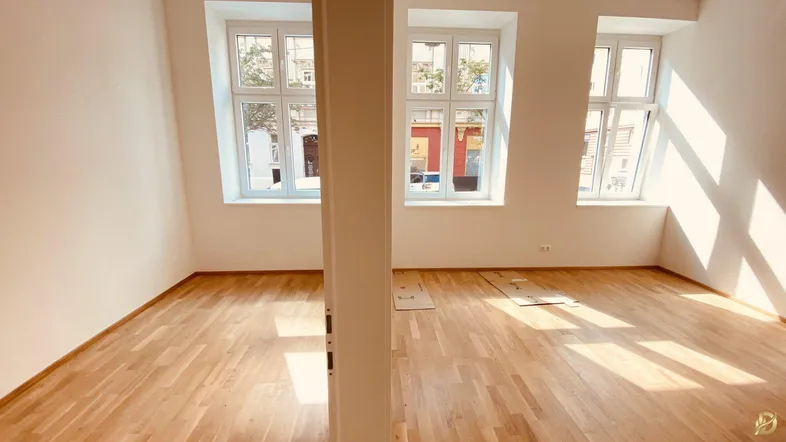 Helles Zimmer mit Holzboden und großen Fenstern, das viel Tageslicht hereinlässt.