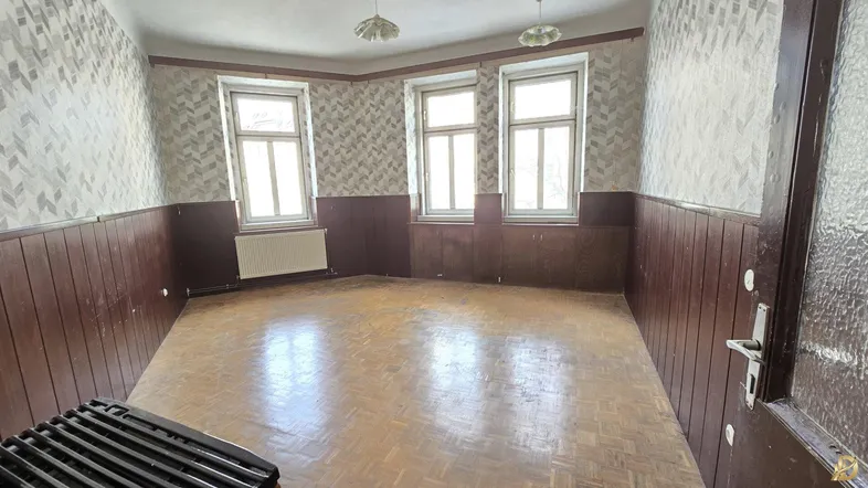 Geräumiges Zimmer mit Fischgrätparkett, Holzvertäfelung und drei großen Fenstern, bereit zur Renovierung.