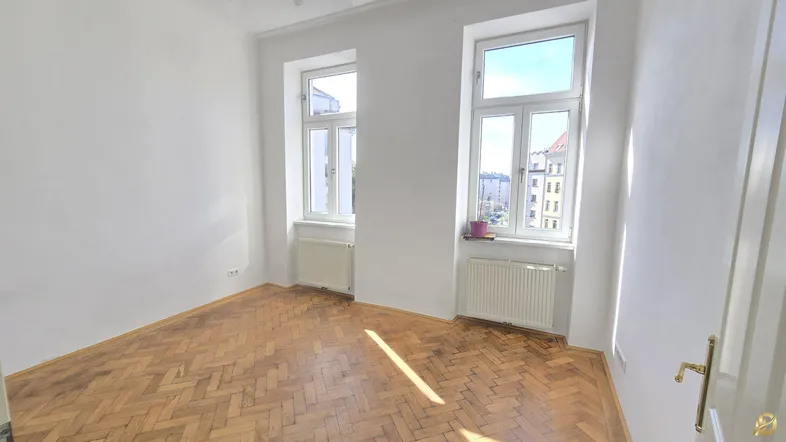 Helles Zimmer mit Fischgrätparkettboden und zwei großen Fenstern, die viel Tageslicht hereinlassen.