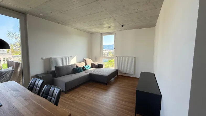 Heller offener Wohn- und Essbereich mit modernem Sofa, Holztisch und großen Fenstern mit Ausblick.