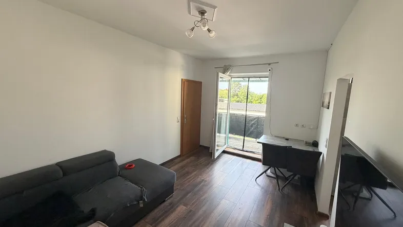 Helles Wohnzimmer mit dunklem Laminatboden, Sofa und Zugang zu einem Balkon.