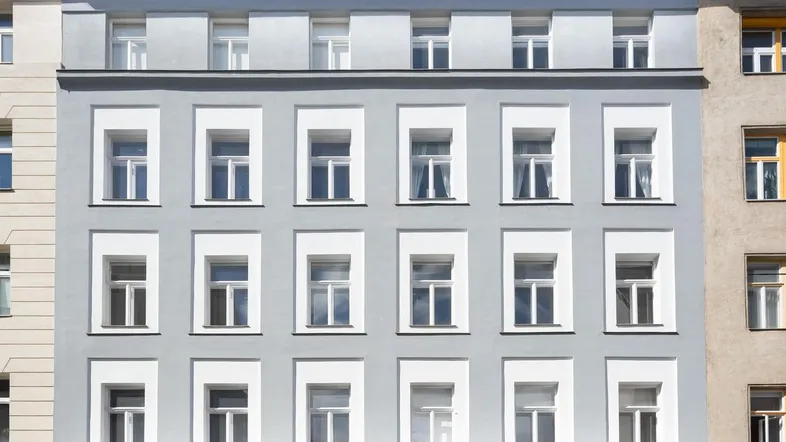 Graue Fassade eines Mehrfamilienhauses mit vielen Fenstern und klarem blauen Himmel.
