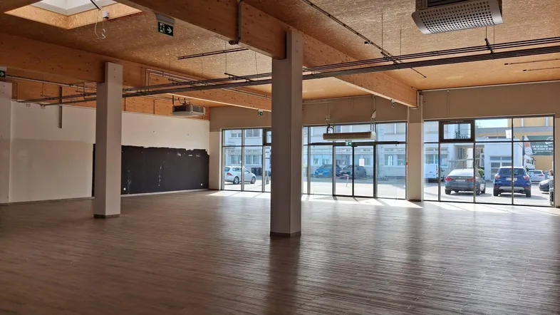 Geräumiger, offener Gewerberaum mit hohen Decken, Holzbalken und großen Fensterfronten für viel Tageslicht.