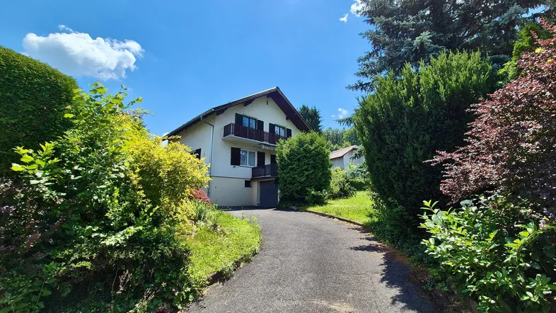 Einfamilienhaus mit Zufahrt und üppiger grüner Umgebung unter blauem Himmel.
