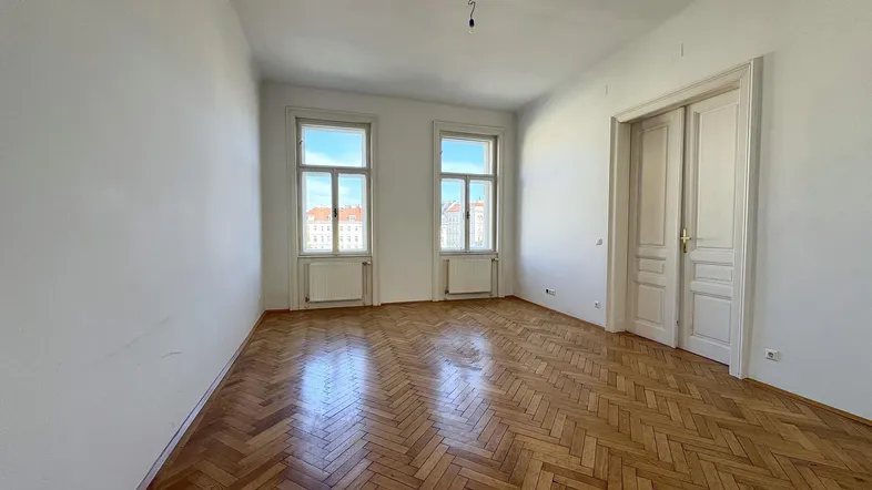 Helles Zimmer mit Fischgrätparkettboden, hohen Decken und großen Fenstern, ideal als Wohn- oder Schlafzimmer.