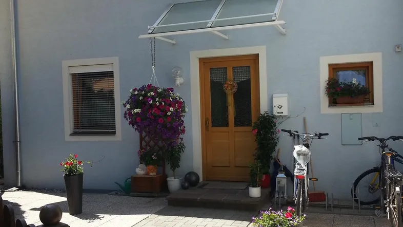 Die helle Hausfassade mit Holztür und Vordach wird von Blumen und Fahrrädern geschmückt.