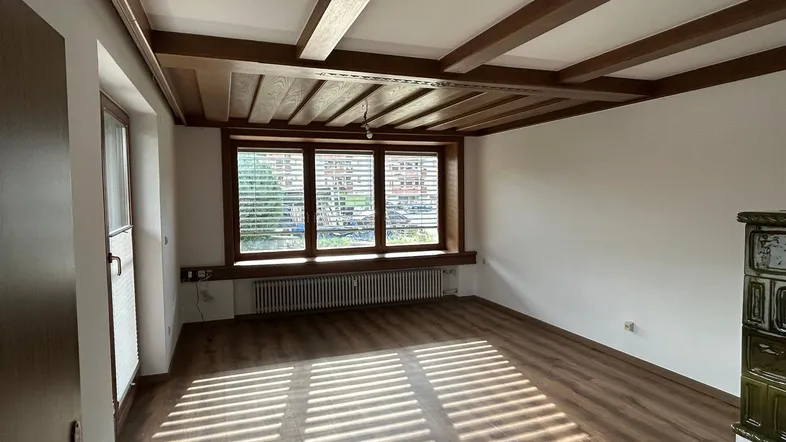 Helles Wohnzimmer mit Holzbalkendecke, Laminatboden und traditionellem Kachelofen.