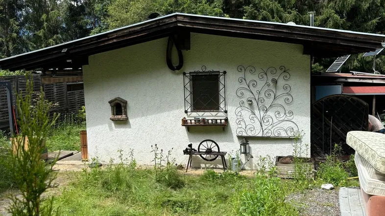 Kleines weißes Gebäude mit dunklem Dach, umgeben von einem verwilderten Garten und hohen Bäumen.