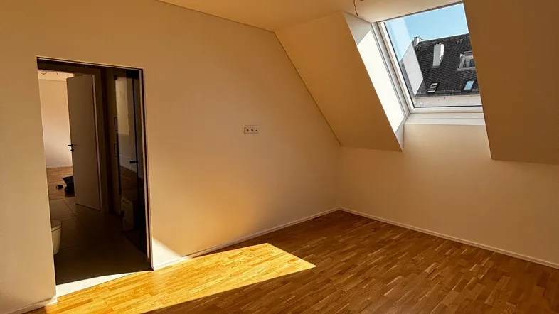 Helles Zimmer im Dachgeschoss mit Holzboden und Dachfenster, durch das Sonnenlicht einfällt.