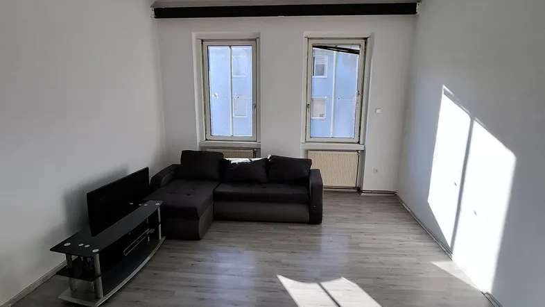Helles Wohnzimmer mit Laminatboden, einem Sofa und Blick aus zwei Fenstern.