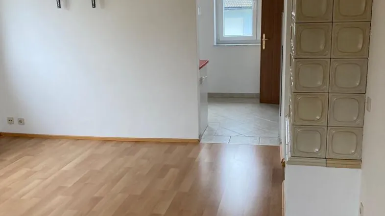 Helles Wohnzimmer mit Laminatboden, einem traditionellen Kachelofen und einem Durchgang zu einem weiteren Raum.