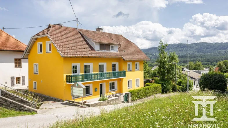Gelbes Haus mit grünem Balkon und Ziegeldach, umgeben von grüner Landschaft und Hügeln.