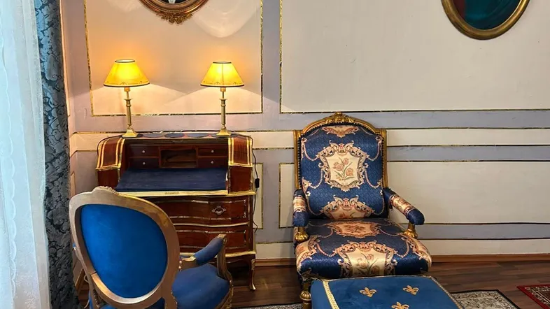 Elegantes Wohnzimmer mit blauen Samtsesseln, antikem Schreibtisch und opulenten Wanddekorationen.