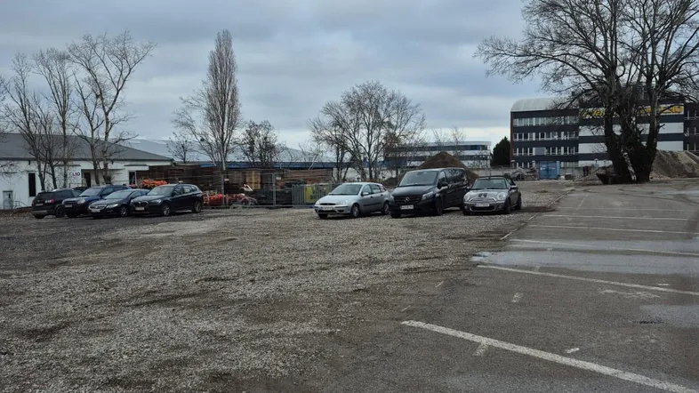Parkplatz im Freien mit geparkten Autos und einem Gebäude im Hintergrund unter bewölktem Himmel.