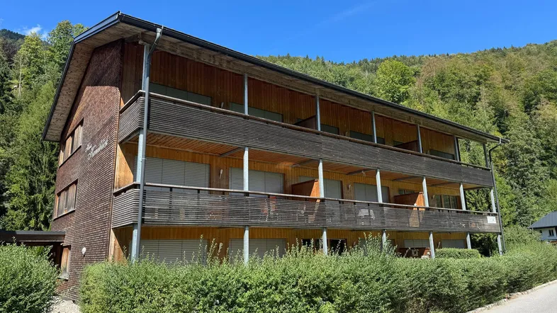 Mehrstöckiges Gebäude mit Holzfassade und Balkonen, umgeben von grüner Natur und Bergen.