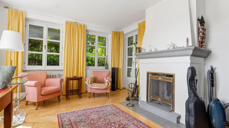 Helles Wohnzimmer mit Parkettboden, Kamin und zwei gemütlichen rosa Sesseln. Gelbe Vorhänge schmücken die Fenster.