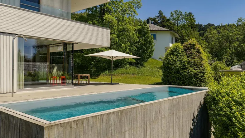 Moderne Villa mit Infinity-Pool und Sonnenterrasse, umgeben von grüner Landschaft und Bäumen.