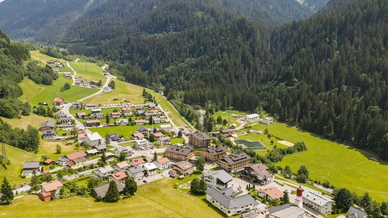 Luftaufnahme eines Dorfes in einem grünen Tal, umgeben von bewaldeten Bergen und Sportanlagen.