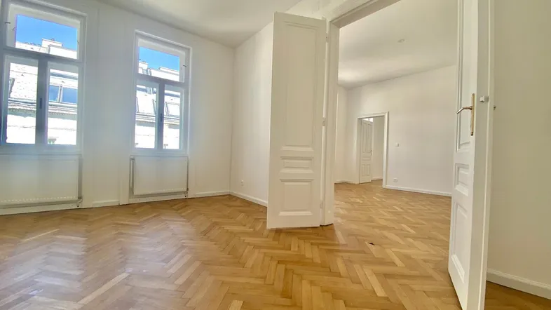 Helles Zimmer mit Fischgrätparkettboden und hohen weißen Türen, das viel Tageslicht hereinlässt.