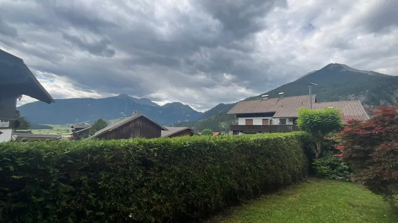 Blick auf die umliegende Berglandschaft mit grüner Hecke und Rasen im Vordergrund unter bewölktem Himmel.
