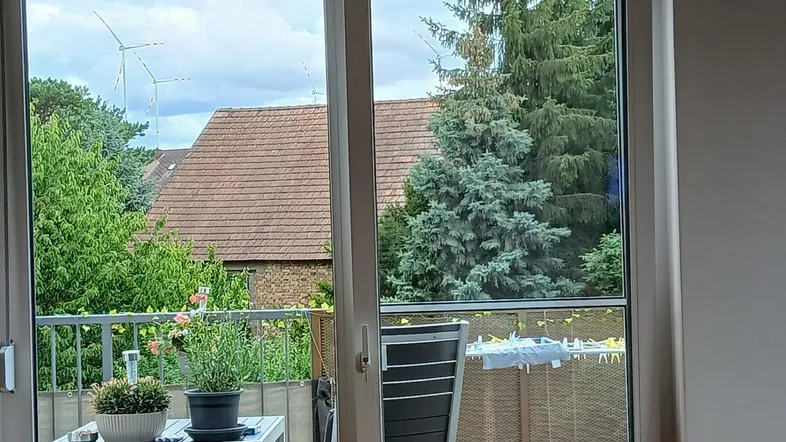 Blick aus einem hellen Raum auf einen möblierten Balkon mit grüner Umgebung und Häusern.