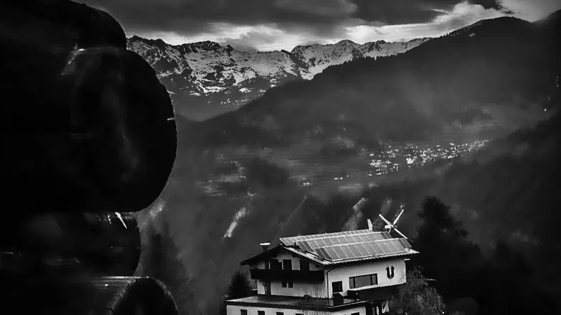 Alpines Haus mit Solaranlage und Windrad, eingebettet in eine dramatische Berglandschaft unter bewölktem Himmel.
