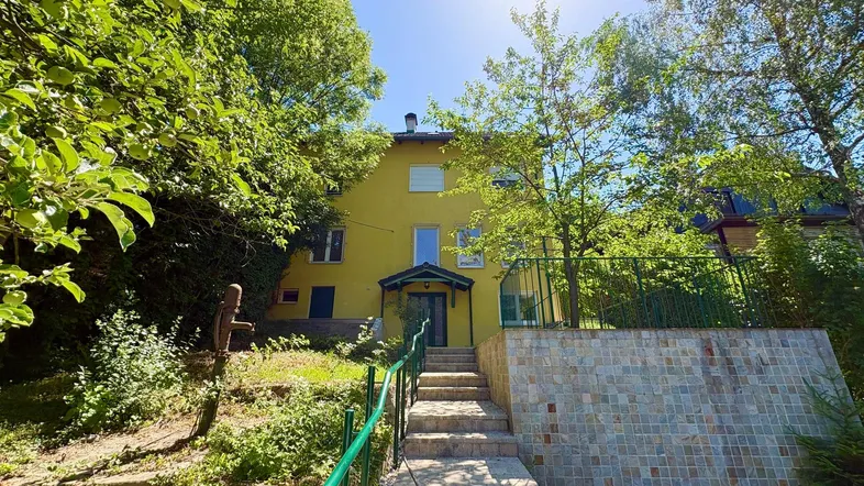 Gelbes Haus mit Treppenzugang und üppiger grüner Umgebung unter strahlendem Sonnenschein.