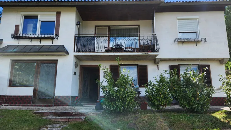 Weißes Einfamilienhaus mit Balkon, Fensterläden und gepflegtem Vorgarten unter blauem Himmel.