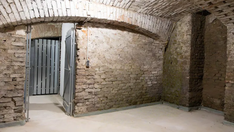 Historischer Kellerraum mit Ziegelgewölbe, unverputzten Wänden und neuem Betonboden, durch eine Metalltür zugänglich.