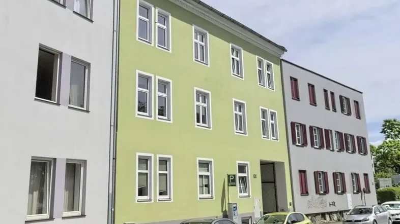 Ansicht der Gebäudefassade mit hellgrünem Anstrich und weißen Fenstern an einer Straße.