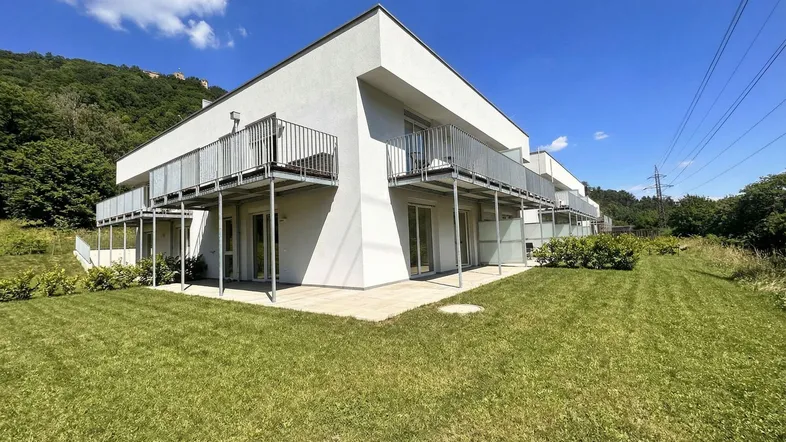 Modernes Wohngebäude mit mehreren Balkonen und einer gepflegten Rasenfläche vor einer grünen Hügellandschaft.