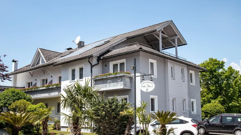 Modernes Gebäude mit Balkonen, Palmen und einem gepflasterten Eingangsbereich unter blauem Himmel.