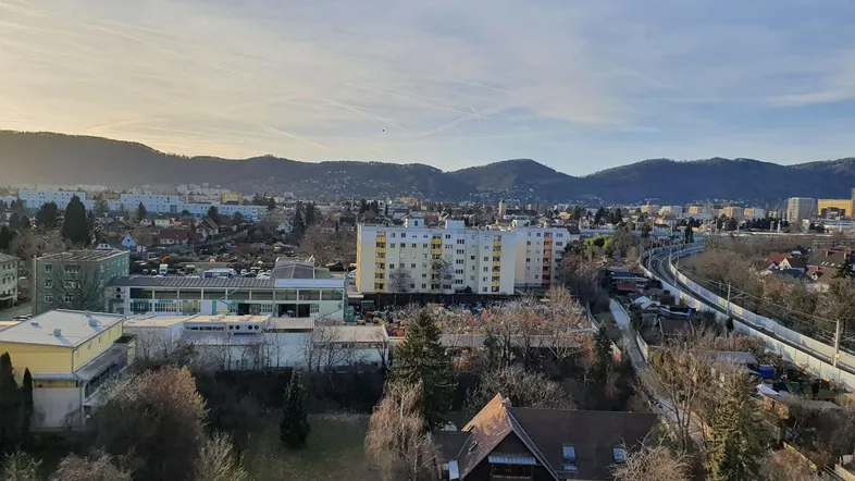 Weitläufiger Blick über die Stadt mit Wohngebäuden, grünen Flächen und Bergen im Hintergrund.