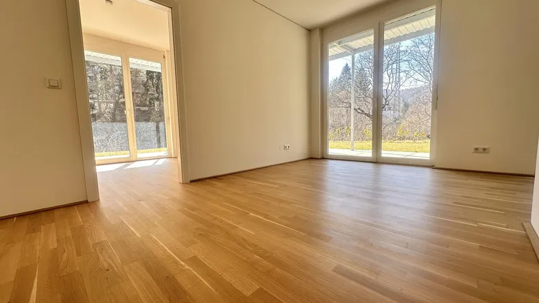 Heller, leerer Raum mit Holzboden und großen Fenstern mit Blick ins Grüne.