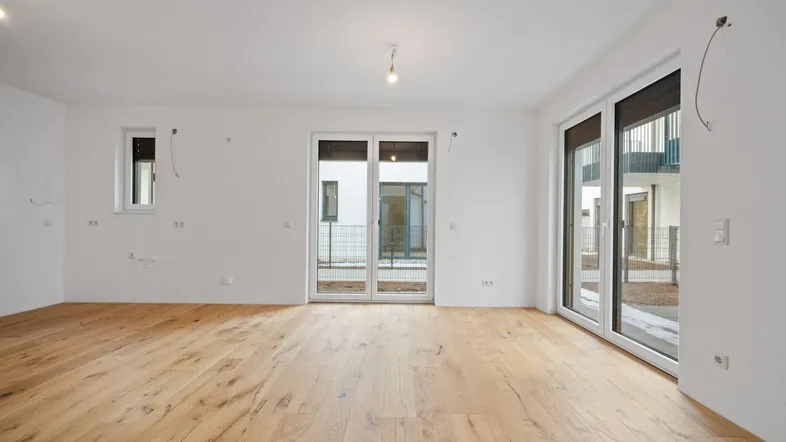 Heller, leerer Raum mit Holzboden und großen Fenstertüren, die ins Freie führen.