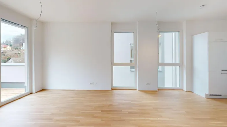Helles, leeres Zimmer mit Holzboden, großen Fenstern und Zugang zu einem Balkon oder einer Terrasse.