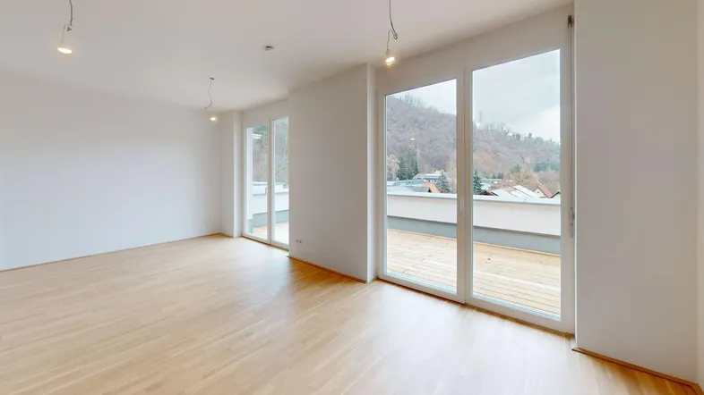 Heller, leerer Raum mit Holzboden und großen Fenstern, die auf eine Terrasse mit Bergblick führen.