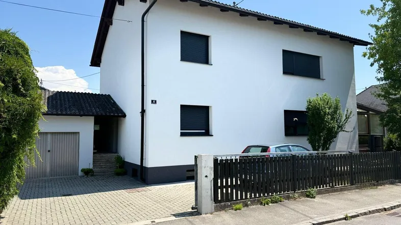 Weißes Einfamilienhaus mit dunklen Fensterläden, gepflasterter Auffahrt und integrierter Garage.