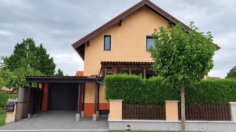 Einfamilienhaus mit orangefarbener Fassade, Carport und gepflegtem Vorgarten unter bewölktem Himmel.