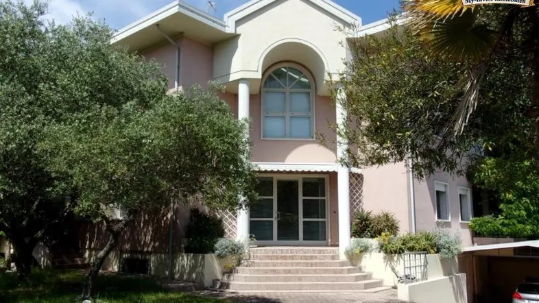 Frontansicht einer eleganten Villa mit gepflegtem Vorgarten, Auffahrt und mediterranen Bäumen.