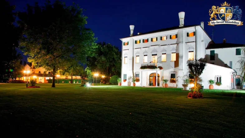 Großes, beleuchtetes Gebäude bei Nacht mit weitläufigem Rasen und stimmungsvoller Gartenbeleuchtung.