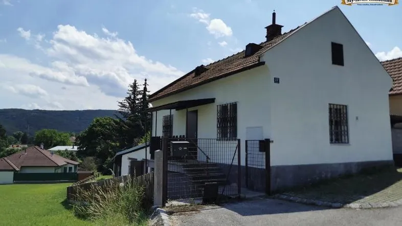 Weißes Einfamilienhaus mit Satteldach und Vorgarten unter blauem Himmel in ländlicher Umgebung.