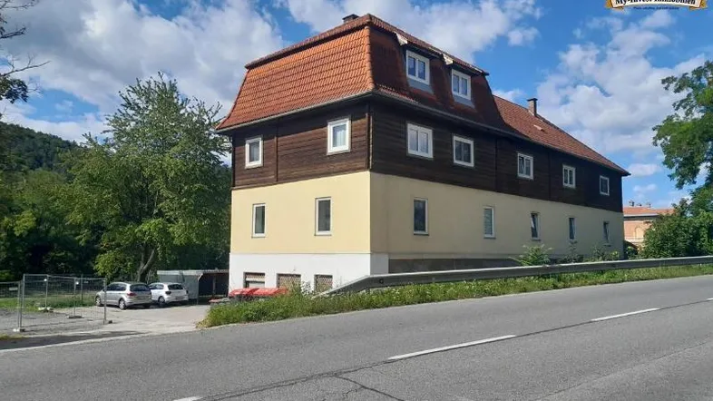 Mehrstöckiges Gebäude mit markantem Mansarddach und Holzverkleidung, umgeben von Bäumen und einer Straße.