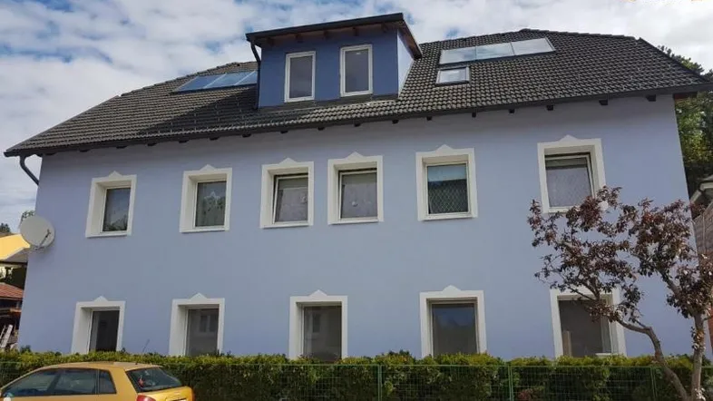 Blau gestrichenes Mehrfamilienhaus mit dunklem Dach und mehreren Fenstern. Ein gelbes Auto parkt davor.