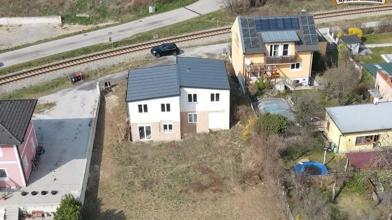 Luftaufnahme des Neubaus mit umliegenden Häusern und Bahngleisen in ländlicher Umgebung.