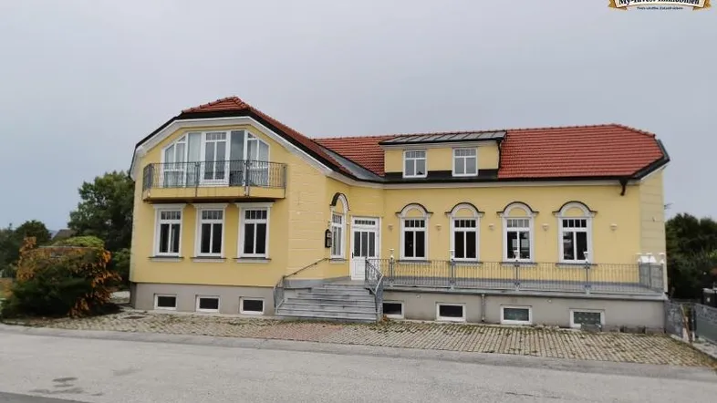 Gelbe Villa mit rotem Ziegeldach, Balkon und Treppenaufgang, umgeben von einer ruhigen Straße.