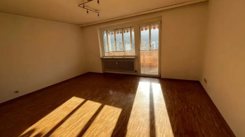 Helles, leeres Zimmer mit Parkettboden und Zugang zu einem verglasten Balkon, durchflutet von Sonnenlicht.
