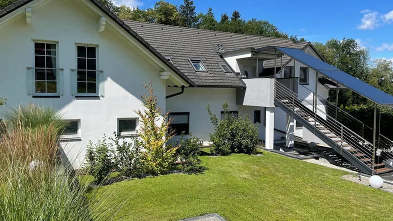 Weißes Einfamilienhaus mit Satteldach, gepflegtem Garten und Außentreppe unter blauem Himmel.